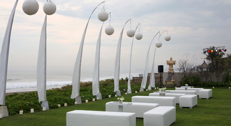 Villa Phalosa — white lounge seating and tall penjor on the lawn above the beach · Seminyak, Indonesia
