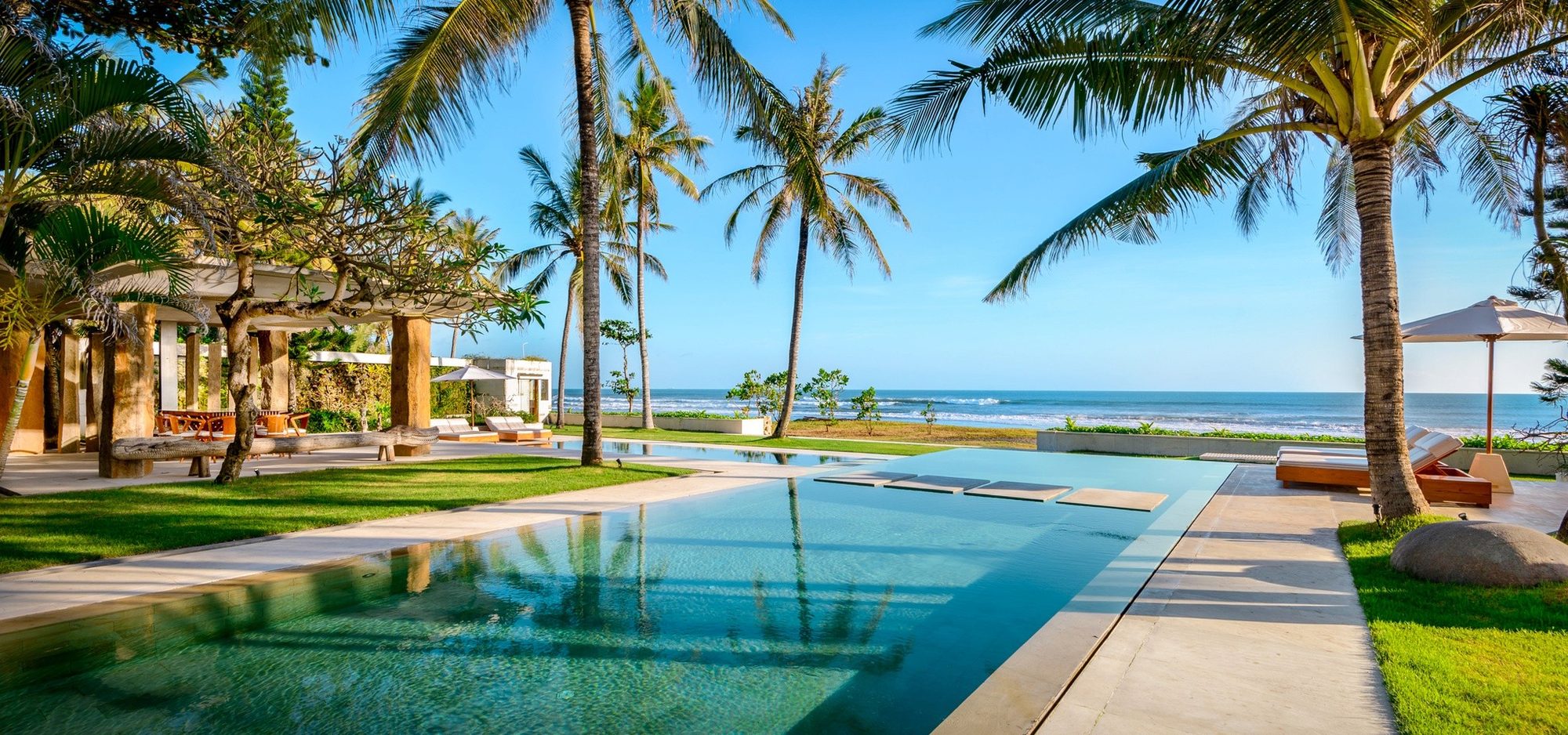 Villa Vedas — the pool and lawn beneath palms, the beach beyond · Bali, Indonesia